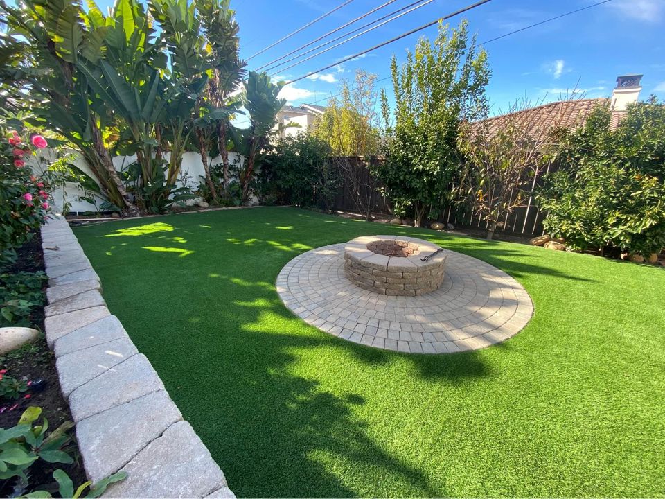 A backyard with artificial grass, bordered by lush green plants and trees. In the center is a circular stone fire pit surrounded by a paved stone patio. The sky is clear and blue.