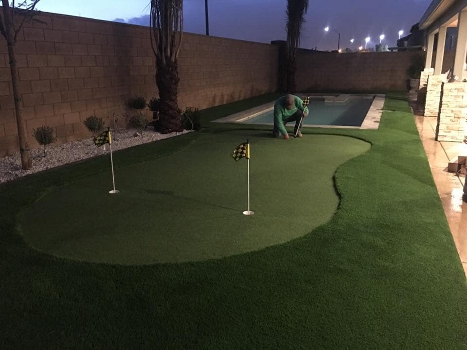 A person is installing or tending to a small artificial putting green with two golf holes and checkered flags in a backyard next to a narrow swimming pool at dusk.