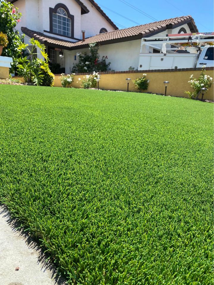 A neatly maintained front yard with lush, green artificial grass, bordered by a yellow wall and flowering plants, in front of a two-story house with a tiled roof and white walls.