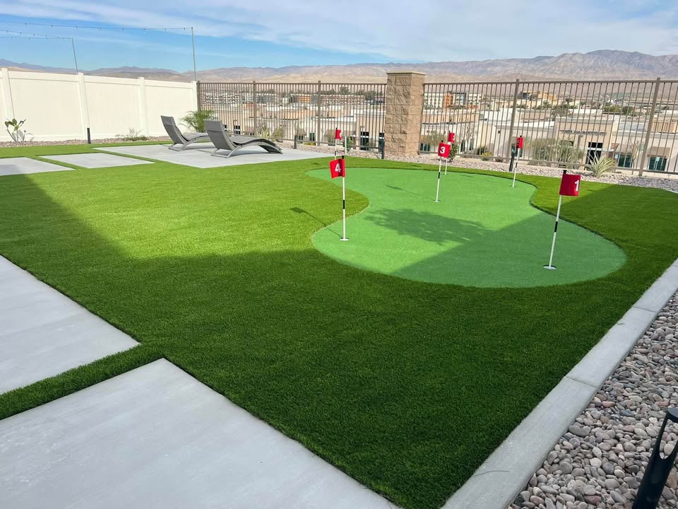A backyard with artificial grass, a small putting green with red flags, lounge chairs on a concrete pad, decorative rocks, and a fence overlooking a city and mountains in the background.