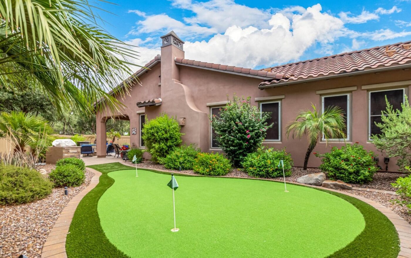 A backyard with a southwestern-style house, manicured landscaping, and stylish putting greens featuring three golf flags, surrounded by rocks, palm trees, and shrubs under a partly cloudy sky.