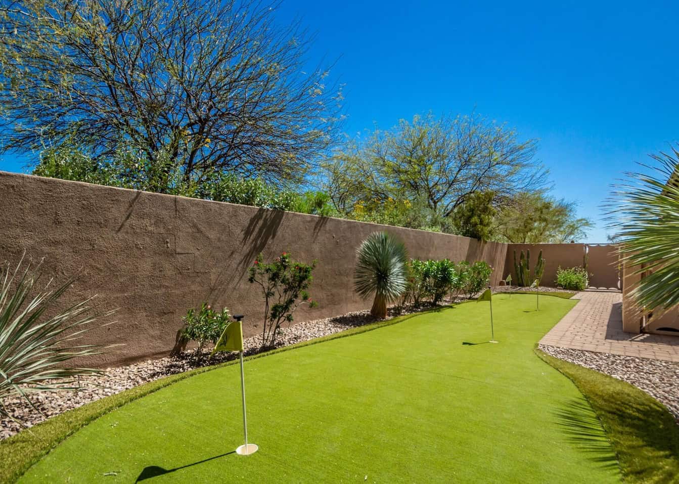 A backyard with custom putting greens, surrounded by desert plants, gravel, and a brown stucco wall under a bright blue sky. Three golf flags are positioned on the green.