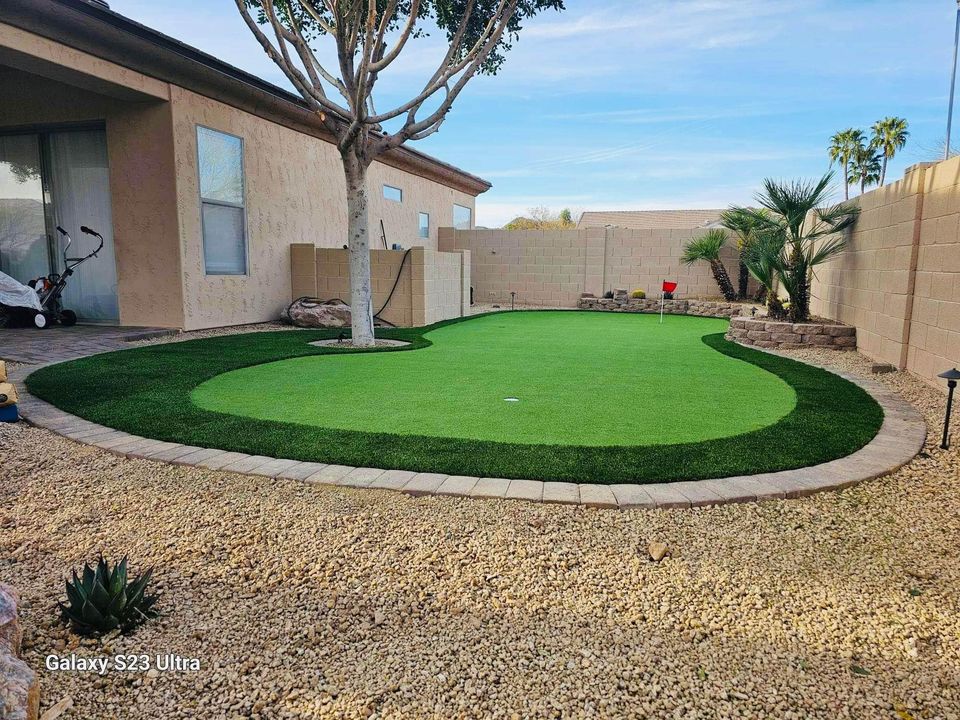 A backyard featuring putting greens with dark artificial turf, bordered by paving stones and gravel, desert plants, and a beige stucco house. A small red flag marks a golf hole on the green.