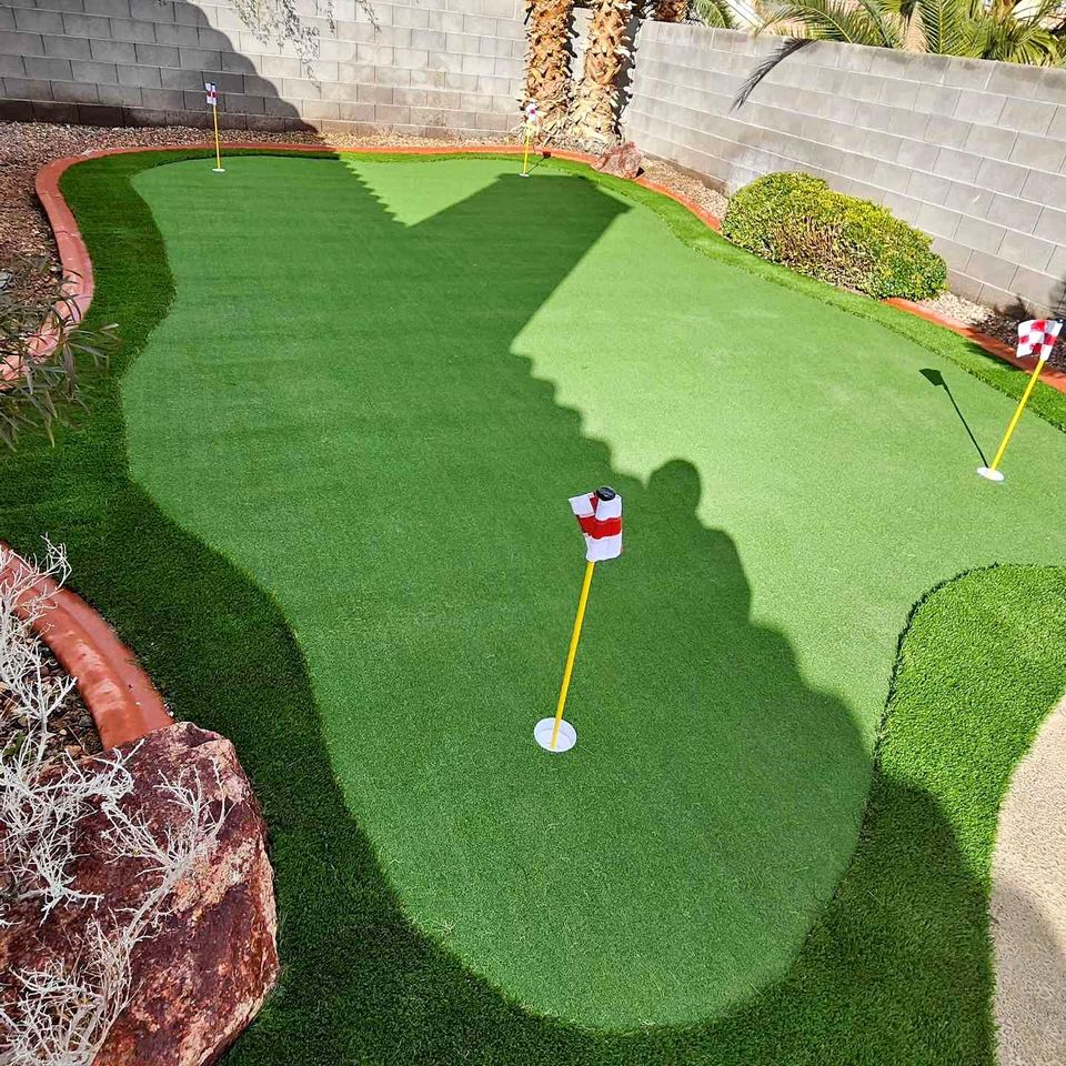 A backyard putting green with synthetic grass, three holes marked by red-and-white checkered flags, bordered by bricks, rocks, and plants—showcasing how modern Putting Greens blend seamlessly with natural elements and dappled shadows from nearby trees.