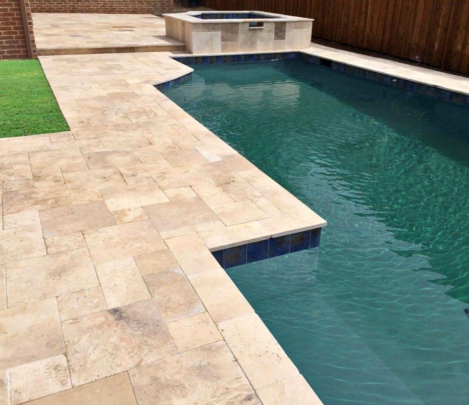 A rectangular swimming pool with light beige stone tiles installed by a skilled pavers installer, adjacent to a small patch of artificial green grass. In the background, a raised hot tub sits next to a wooden fence.
