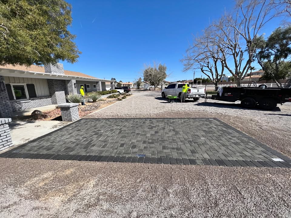 A pavers installer places a new rectangular section of dark gray pavers in the driveway of a suburban home on a clear, sunny day. Construction vehicles and equipment are visible nearby, with trees lining the property.