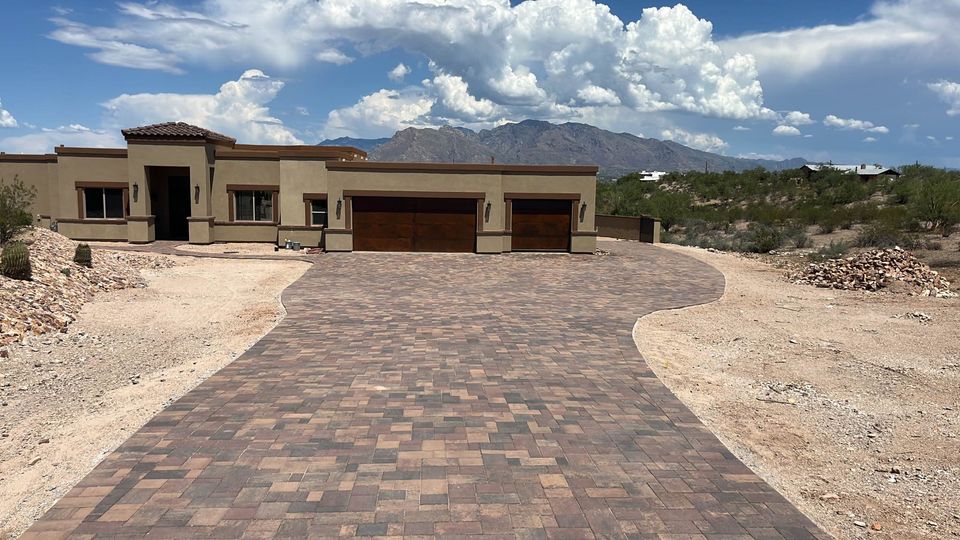 A modern single-story house with a brown tiled driveway crafted by a skilled pavers installer, surrounded by desert landscape and mountains in the background under a partly cloudy sky.