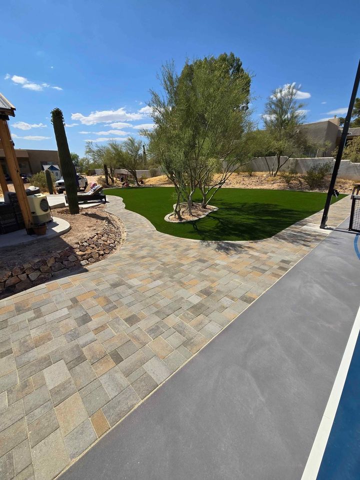 A spacious backyard features a stone patio walkway crafted by a skilled pavers installer, artificial turf with trees and desert landscaping, and a pool edge in the foreground under a sunny, clear blue sky.