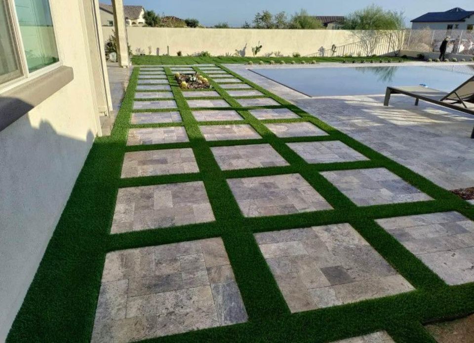 A modern backyard patio features large square stone tiles arranged in a grid with strips of green grass between them, expertly crafted by a skilled pavers installer, next to a swimming pool and lounge chairs.