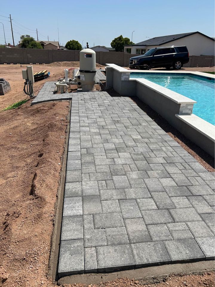 A newly installed gray stone paver walkway by an expert pavers installer runs alongside a rectangular backyard swimming pool, with unfinished landscaping and construction materials nearby. A black SUV is parked in the background.
