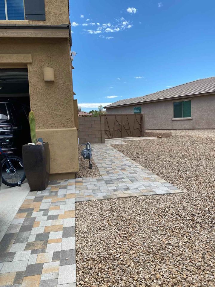 A paved walkway, crafted by a skilled pavers installer, leads from a garage past a potted cactus and through a gravel yard to a gate in a stone wall. The sky is partly cloudy, with another house visible in the background.