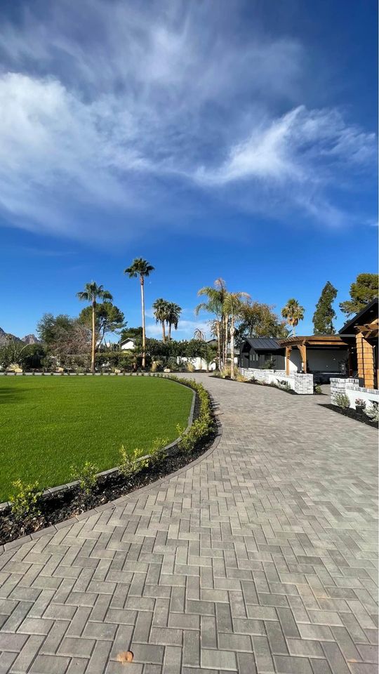 A paved stone pathway, crafted by a skilled pavers installer, curves alongside a green lawn bordered by small shrubs and palm trees under a blue sky. Buildings with wooden and white exteriors stand to the right.