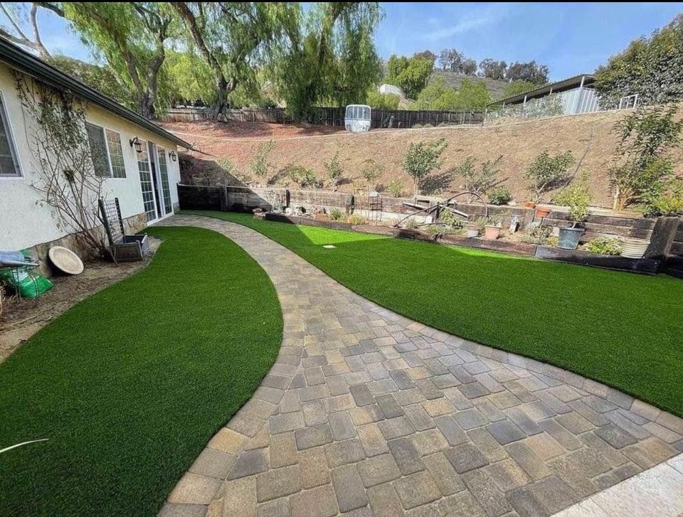 A backyard with a curved stone walkway, crafted by a skilled pavers installer, divides two areas of artificial green grass bordered by a raised garden bed and hillside with scattered plants and trees. A white house sits on the left side of the image.