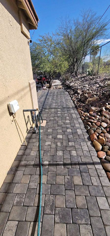 A paved walkway, crafted by a skilled pavers installer, runs beside a tan house and is bordered by rocks and desert plants beneath a clear blue sky. A green garden hose lies along the walkway.