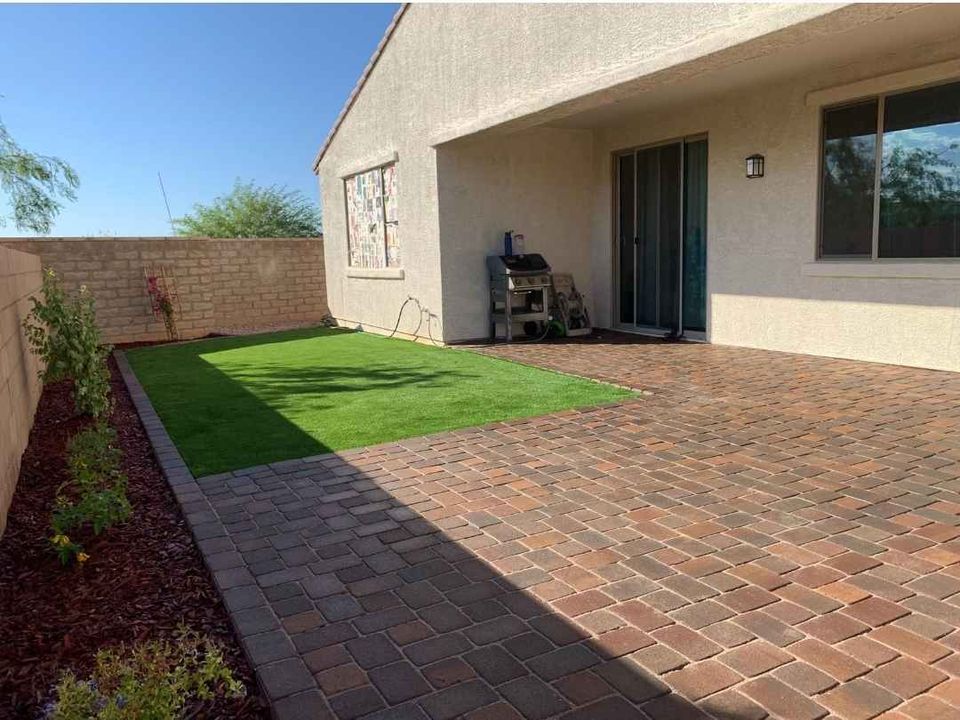 A backyard patio with professionally laid brick pavers by a skilled pavers installer, a small square patch of artificial grass, a barbecue grill near the sliding glass door, and a garden bed with plants along the wall.